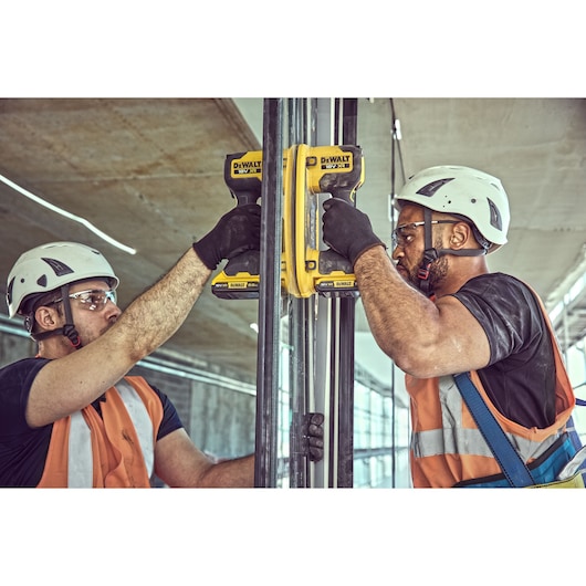 Two workers using DEWALT DCE590 tools on metal pipes at a construction site, wearing safety helmets and vests.
