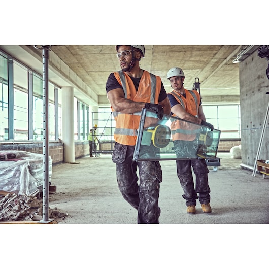 Two workers in safety gear carry glass with suction tools at a construction site.