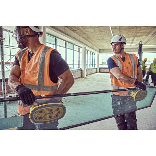 Two workers use DEWALT suction tools to carry glass indoors, both wearing safety gear and orange vests.