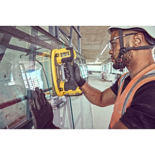 Person uses DEWALT DCE590 tool to lift glass at construction site, wearing gloves and safety gear.