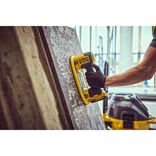 A DEWALT cordless tool is used to sand a large board. Worker wears gloves. Workshop environment.