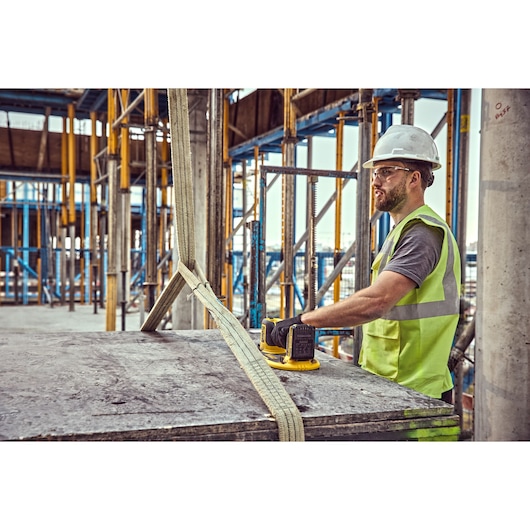 Worker in safety gear uses DEWALT tool on construction site, holding a device on a concrete slab with straps.