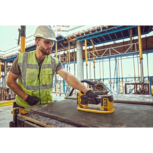 Worker uses DEWALT DCE590 tool on a construction site, wearing safety gear and gloves.