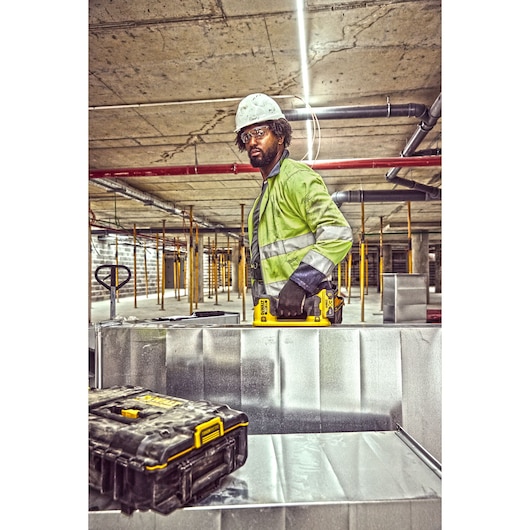 Worker in safety gear holds a DEWALT DCE590 tool at a construction site. DEWALT case is visible in foreground.