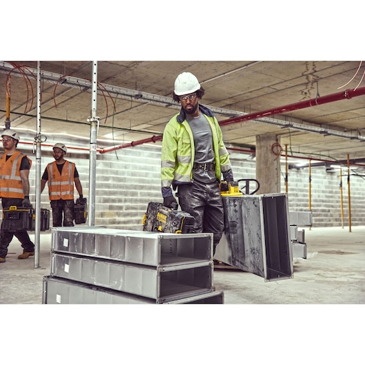 Construction workers carry DEWALT toolboxes and metal parts at an indoor job site with unfinished concrete and scaffolding.