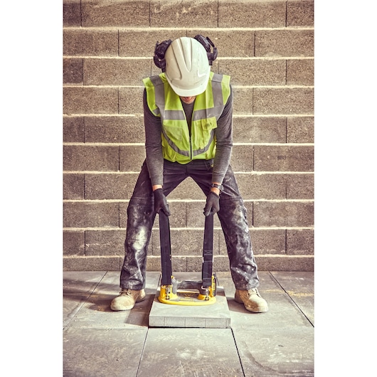 Worker uses DEWALT DCE590 to lift paving slab, wearing safety gear and gloves, with brick wall background.