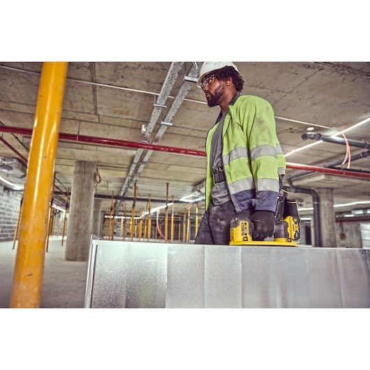 Worker carries metal sheet using DEWALT DCE590 tool in a construction site, wearing safety gear and gloves.