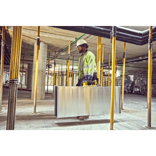 Worker in safety gear carries a metal case with yellow handles in a construction site with pillars.