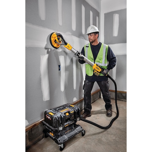 A construction worker wearing a hard hat and safety vest is sanding a drywall using a DEWALT cordless drywall sander, model DCE800T2. The sander is connected by a hose to a portable dust extractor placed on the floor.
