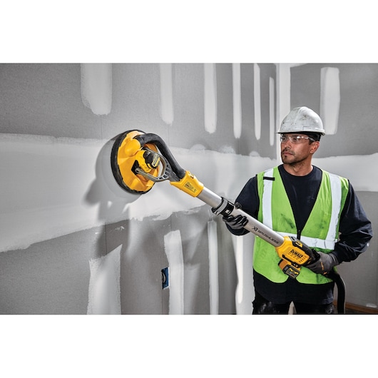 A person wearing a hard hat and a safety vest is using a DEWALT DCE800T2 cordless drywall sander on a wall with unfinished plaster joints.