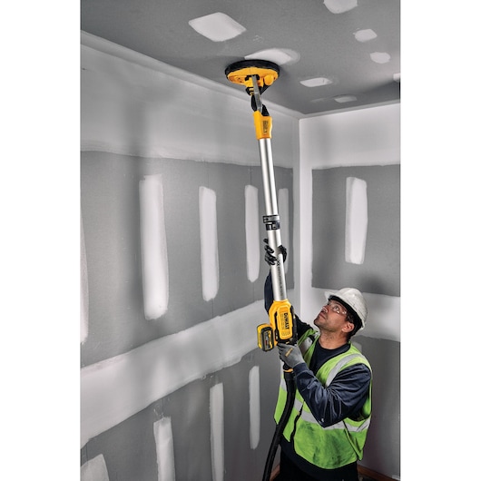 A worker in a safety vest and hard hat uses a DEWALT cordless drywall sander with an extension pole to sand the ceiling in a room with unfinished drywall.