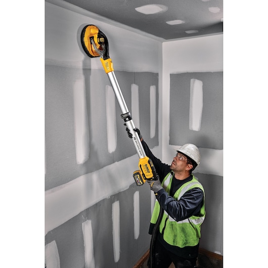 A worker in a safety vest and helmet uses a DEWALT cordless drywall sander to sand a wall and ceiling in a room with unfinished drywall.