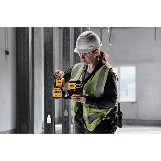 Worker uses DEWALT DCE822D1G18 laser tool at a construction site, wearing safety gear and a reflective vest.