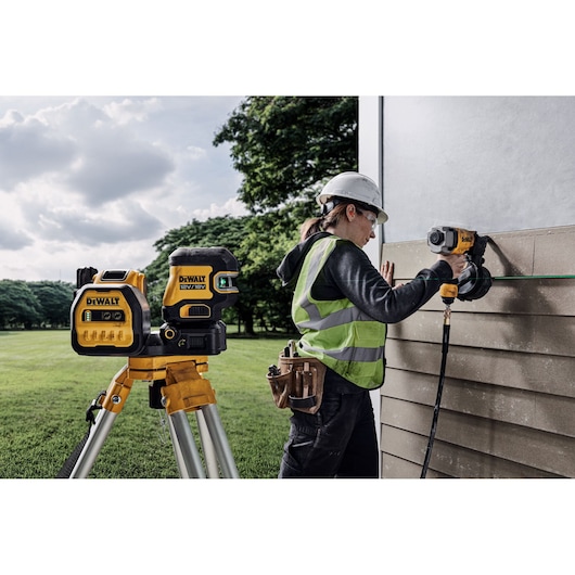 DEWALT laser level on tripod and worker installing siding outdoors with power tool.