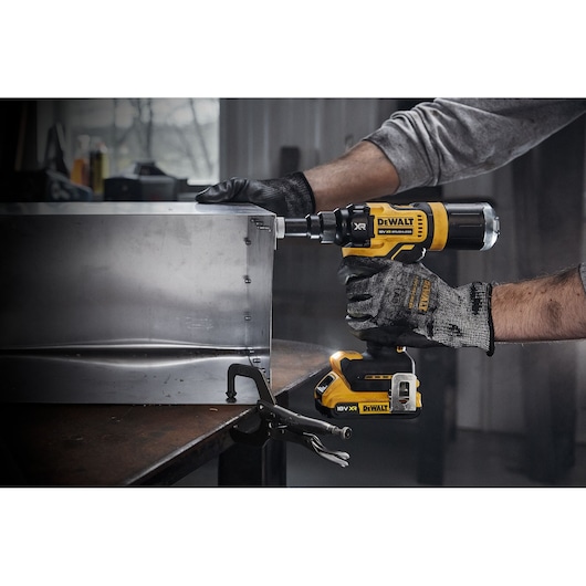 A person wearing work gloves is using a DEWALT DCF403NT cordless power tool to fasten screws into a metal box in a workshop.