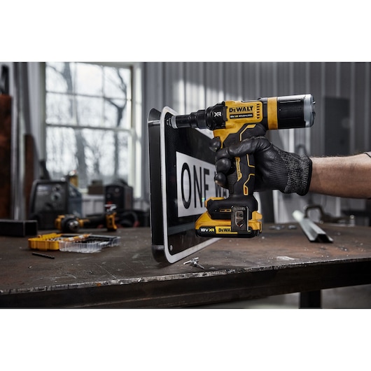A person wearing a black work glove uses a DEWALT DCF403NT 18V XR cordless drill to drill into a metal sign on a workbench in a workshop.
