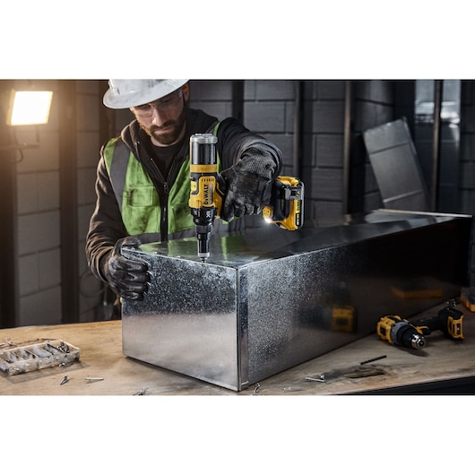 A person wearing safety gear uses a DEWALT DCF403NT cordless power tool to work on a metal box in an industrial workshop. Various screws and tools are visible on the workbench.