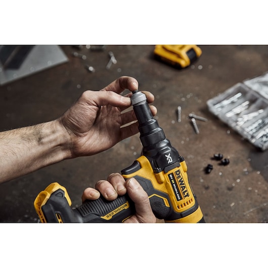 A person is holding a DEWALT cordless power tool, model DCF403NT, on a workbench with various screws and parts scattered around, adjusting the tool's attachment.