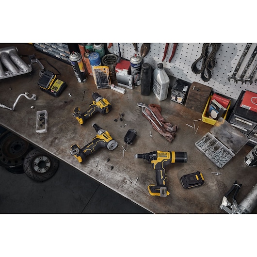 A workbench with several DEWALT power tools, including a cordless ratchet, scattered screws, batteries, cleaning cloth, tools, and other equipment. The workspace is organized with various tools hanging on a wall pegboard.