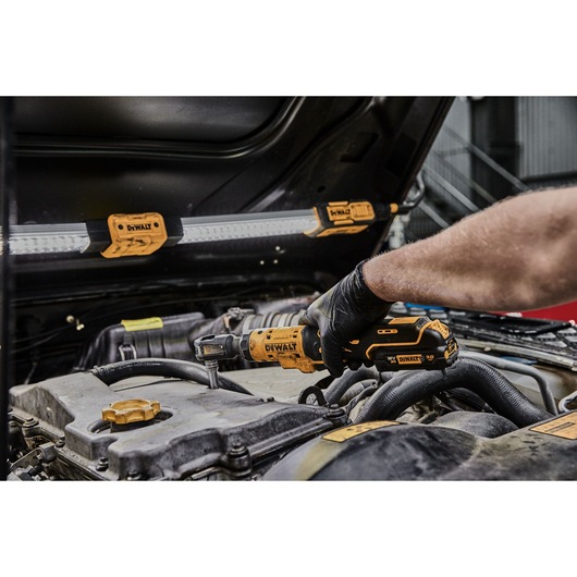 A person wearing a black glove uses a yellow DEWALT cordless ratchet tool (DCF503L1G) to work on an engine under the hood of a car. Several DEWALT LED work lights are attached to the underside of the hood for illumination.