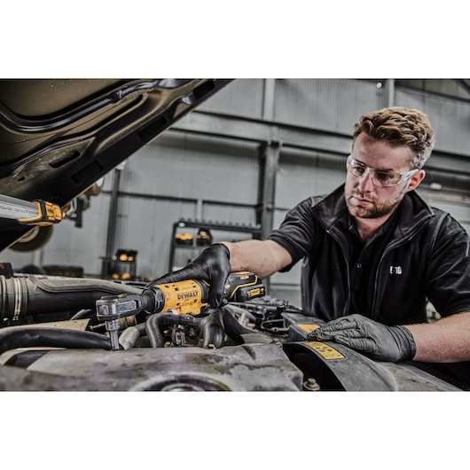 A person wearing black gloves is using a DEWALT DCF503N cordless ratchet tool to work on a car engine. The person's face is blurred, and various engine components and tools are visible in a workshop setting.