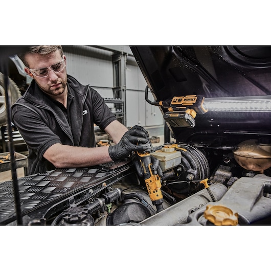 A person wearing black gloves uses a DEWALT DCF503N cordless ratchet wrench to work on a car engine in a garage. There is a DEWALT work light attached to the car hood, illuminating the workspace.