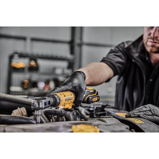 A person wearing black gloves uses a DEWALT DCF504N cordless ratchet tool to work on a car engine in a garage setting.