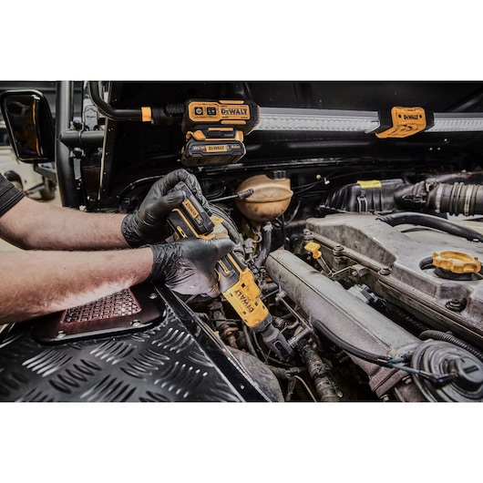 A person wearing black gloves using a DEWALT cordless ratchet tool to work on a car engine in a workshop, with other DEWALT tools visible in the background.