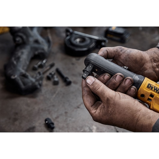 Hands holding a DEWALT cordless ratchet tool with bolts and metal parts visible on a workshop table.