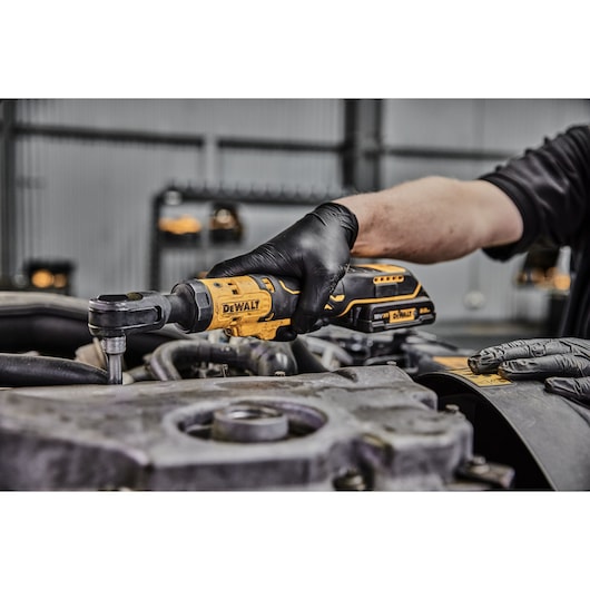 A person wearing black gloves uses a DEWALT cordless ratchet wrench to work on an engine component in an industrial setting.
