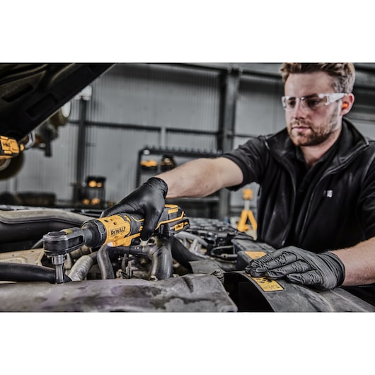 A person wearing black gloves is using a DEWALT cordless ratchet tool (DCF513N) to work on a car engine in a workshop. The