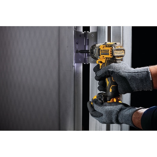 Gloved hands use a DEWALT DCF601N cordless screwdriver to tighten screws on a metal hinge.