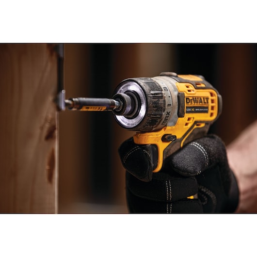 A DEWALT cordless screwdriver being used to drive a screw into wood, held by a gloved hand.