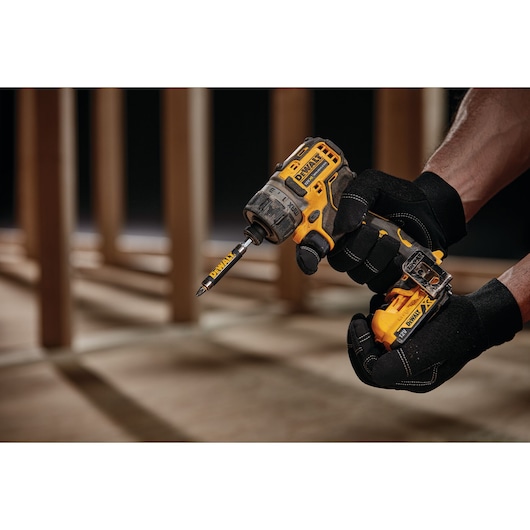 Gloved hand holding a DEWALT cordless screwdriver drill with bit attached, indoors near wooden frames.