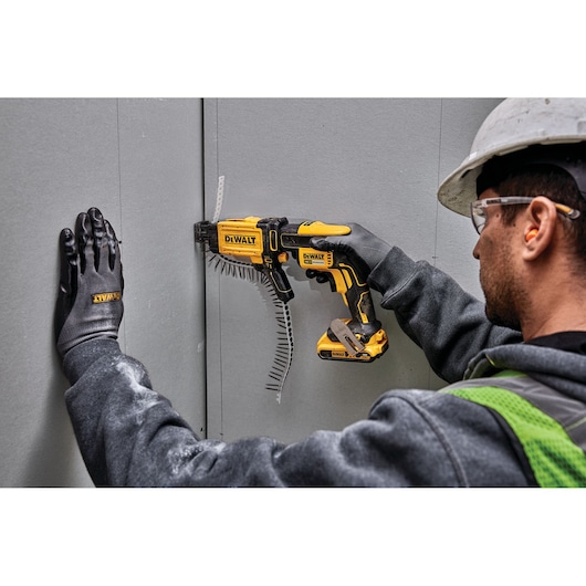 Worker uses DEWALT DCF6202 tool to attach drywall, wearing gloves, helmet, and safety glasses.