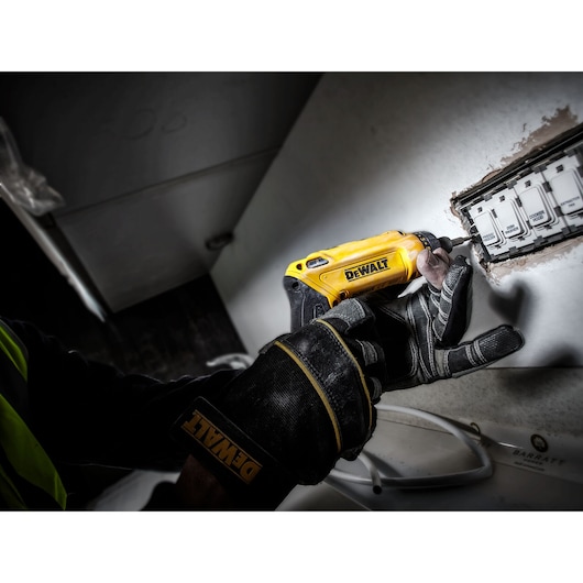 Gloved hand uses DEWALT DCF680G2 screwdriver near an electrical outlet on a wall.