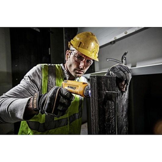 Worker uses DEWALT DCF680G2 tool on a metal surface, wearing gloves, vest, and helmet at a construction site.