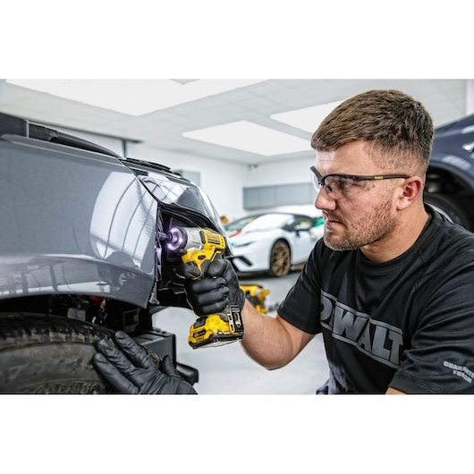 A person wearing black gloves uses a yellow DEWALT cordless drill to work on the front end of a gray car in a garage. The face of the person is blurred for privacy and there are other cars in the background.