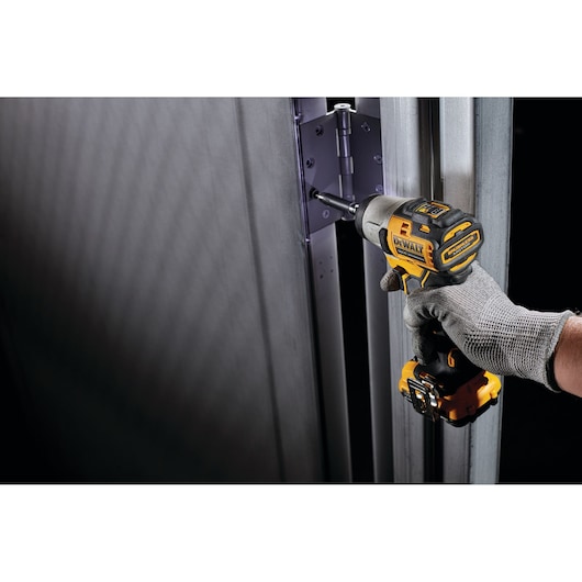 A person wearing a grey work glove uses a DEWALT cordless impact driver to install or tighten screws on a metal door hinge.