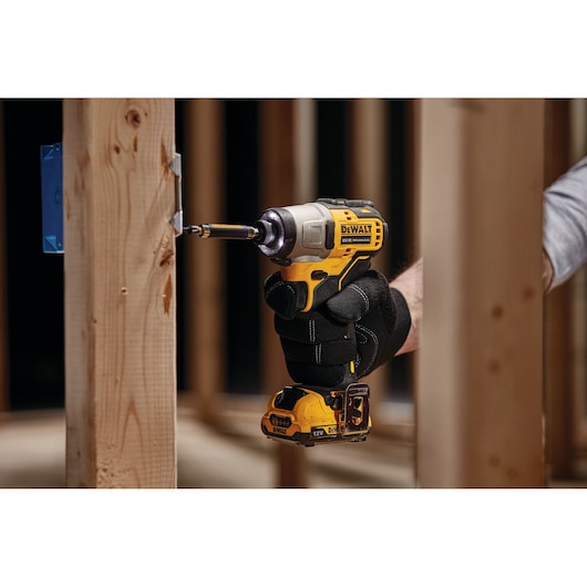 A person wearing black gloves is using a DEWALT cordless impact driver to drive a screw into a wooden frame in a construction setting.