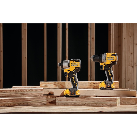 Two DEWALT cordless power tools, including a drill and an impact driver, are placed on stacked wooden planks in a workshop setting with unfinished wood framing in the background.