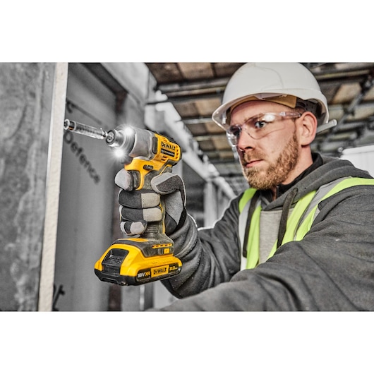 Person wearing a safety helmet and high-visibility vest using a DEWALT DCF840D2T cordless impact driver on a construction site. The person's face is blurred for privacy.