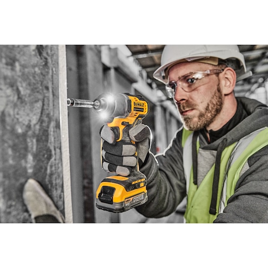 A person wearing safety gear uses a DEWALT DCF840NT cordless impact driver to fasten a screw into a wall at a construction site. The tool is yellow and black, and the person’s face is obscured.