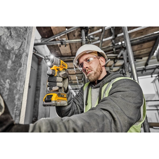 A person wearing safety gear uses a yellow DEWALT cordless drill to fasten a screw into a metal surface on a construction site.