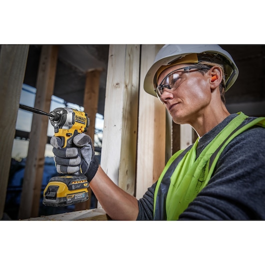 A person wearing a safety helmet, neon safety vest, and gloves uses a DEWALT DCF850E2T 18V cordless impact driver to work on