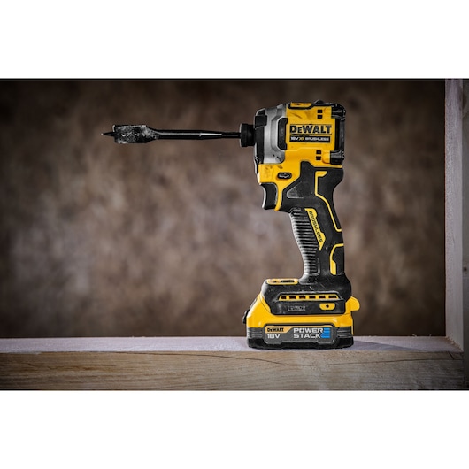 A DEWALT 18V XR brushless cordless impact driver with a black and yellow design. The tool is fitted with a drill bit and features a POWERSTACK battery at the base. It is placed on a wooden surface with a blurred brown background.