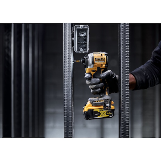 Gloved hand using a DEWALT DCF850NT impact driver to screw into a metal frame near an electrical box.