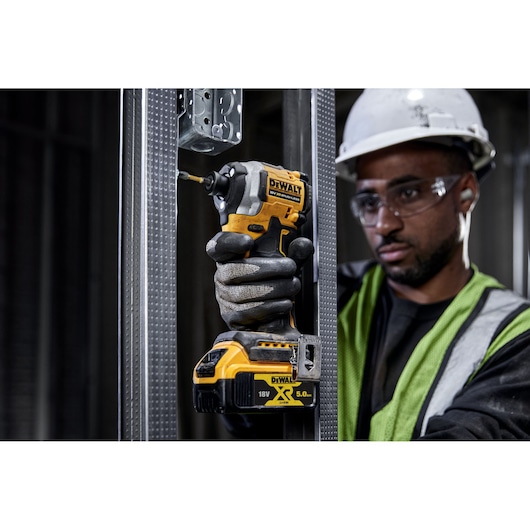 A gloved hand uses a DEWALT DCF850NT impact driver to screw into a metal frame at a construction site.