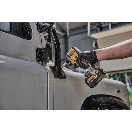 A gloved hand using a DEWALT cordless impact driver on a truck door hinge.