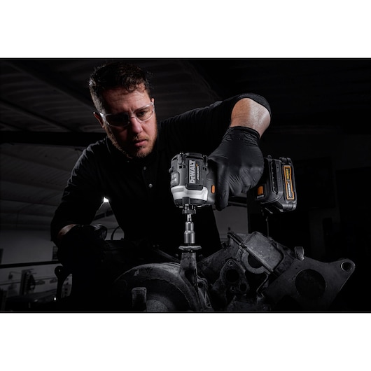 A person with a blurred face uses a DEWALT cordless tool, model DCF85ME2GT, to work on mechanical equipment. The person is wearing black gloves and is in a workshop setting.